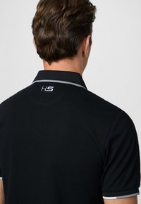 Hombre con cabello castaño que lleva una camiseta de polo negra con ribete gris en el cuello y las mangas, vista trasera mostrando el logo "HS" debajo del cuello.