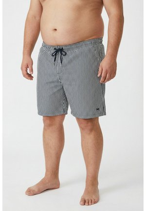 Mężczyzna w czarno-białych, pionowo paskowanych kąpielówkach ze sznurkiem, stojący boso na białym tle.