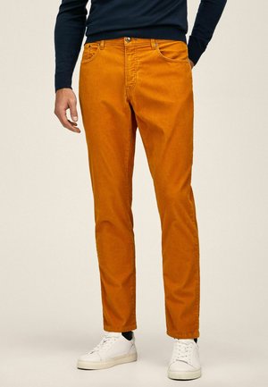 Man wearing slim-fit orange pants, white sneakers, and a navy long-sleeve top, standing with one hand in pocket.