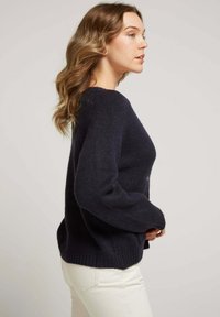 Pull en tricot bleu marine avec une coupe décontractée, ourlet et poignets à côtes, doté de longues manches et d'un tissu doux et texturé. Associé avec un jean clair.