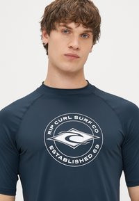Marinblå rash guard i elastiskt material, med en rund vit logotyp som har texten "Rip Curl Surf Co" och "Established 69". Kortärmad.