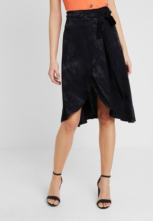 Femme portant une jupe portefeuille noire avec une ceinture à nouer, associée à des talons noirs à bride cheville et un top côtelé orange avec un bouton.