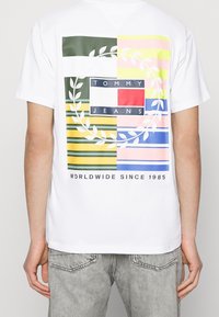 Witte katoenen t-shirt met een gekleurde grafische print op de achterkant, met strepen en een laurierkrans ontwerp, gelabeld "TOMMY JEANS" en het jaar 1985.