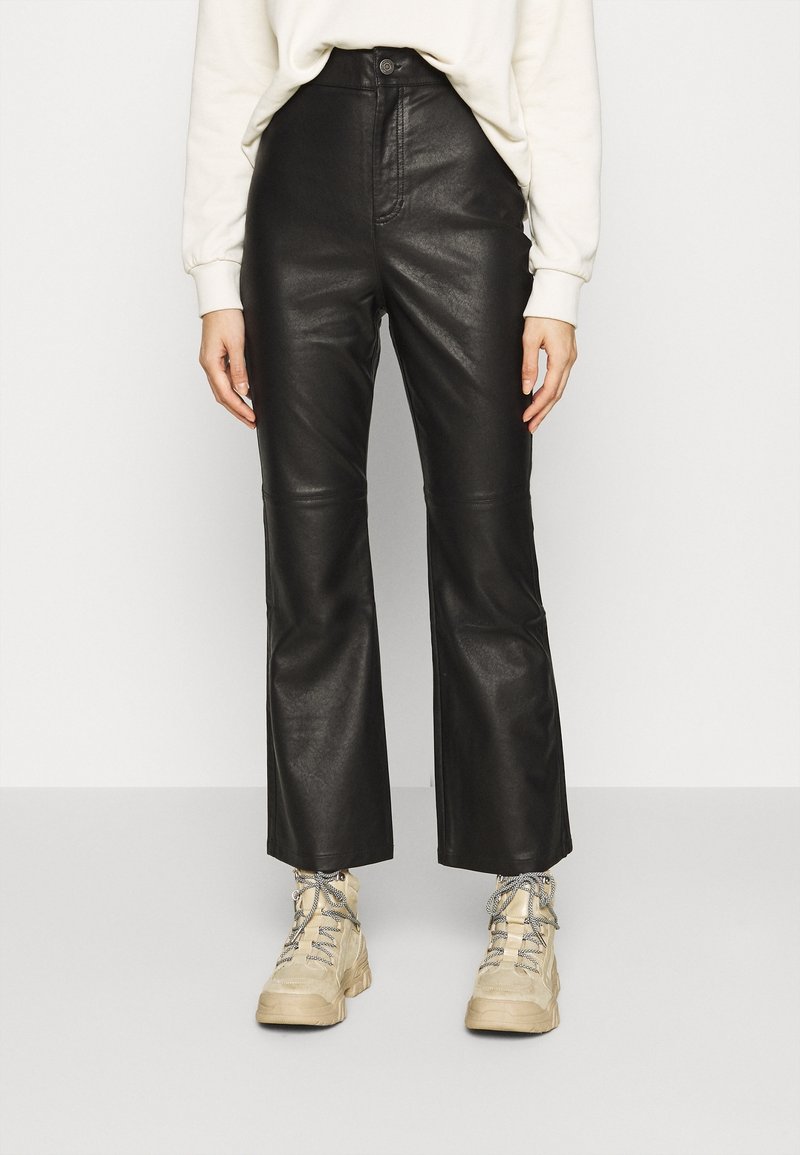 Pantalon évasé en similicuir noir à taille haute, avec une fermeture à boutons sur le devant et des détails de couture sur les côtés. Porté avec des bottines beiges.