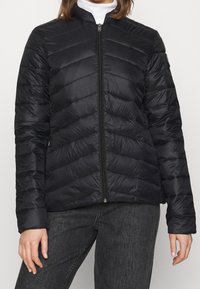 Svart quiltad pufferjacka med hög krage, dragkedja och snedställd chevronmönster på tyget. Slätt, lätt material.