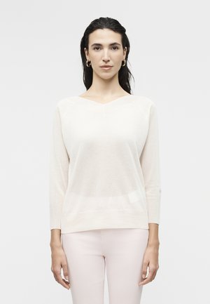 Vrouw met donker haar draagt een crème V-hals trui en lichtroze broek, staand tegen een effen lichte achtergrond.