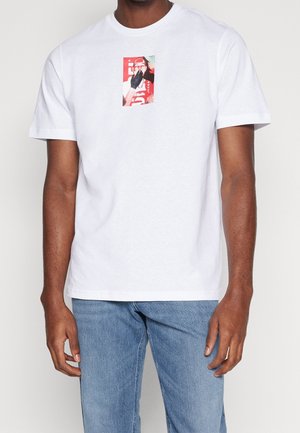 Uomo che indossa una t-shirt bianca a maniche corte con una piccola grafica multicolore sul petto e jeans azzurri chiari su sfondo uniforme.
