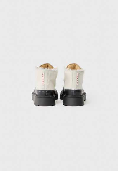 Ankelboots med en krämfärgad päroverdel, svart gummisula och röd märkessymbol på baksidan, med en texturerad yta.