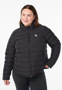 Frau, die eine schwarze Daunenjacke mit Reißverschluss und Logo trägt, steht mit der Hand in der Tasche vor einem weißen Hintergrund.