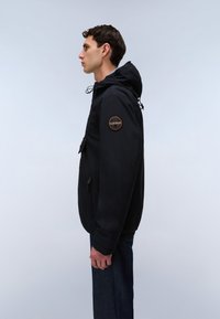Nuori mies, jolla on musta huppari ja pyöreä logopatch yläkädessä, seisoo sivuprofiilissa yksiväristä taustaa vasten.