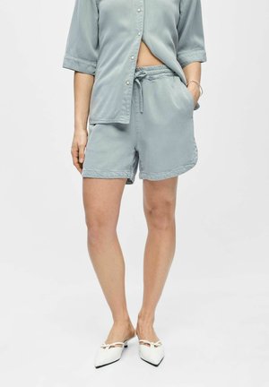 Persona con camisa azul claro de botones y pantalones cortos a juego con cintura elástica, combinados con zapatos blancos de tacón bajo y punta puntiaguda.