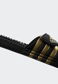 Svart og gull slide sandaler med en teksturert stropp og tre markante striper. Har teksturert såle og merkevare langs siden.