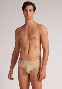 Beige Herren-Brief aus glattem Stoff mit schmaler Passform, breitem Bund und dezenten Nähten, die einen tief sitzenden Schnitt bieten.