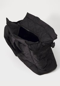 Sac matelassé noir au design structuré, doté d'une fermeture éclair sur le dessus et d'une bandoulière réglable. L'intérieur est ouvert, mettant en valeur une texture lisse.