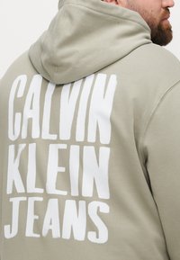 Oliwkowa bluza z kapturem z białym, pogrubionym napisem "CALVIN KLEIN JEANS" na plecach, z kapturem ze sznurkiem i miękką fakturą materiału.