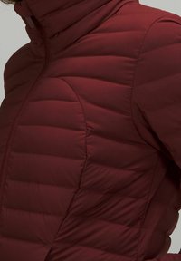 Zbliżenie na osobę ubraną w bordową pikowaną kurtkę puchową z wysokim kołnierzem i zamkiem błyskawicznym z przodu.