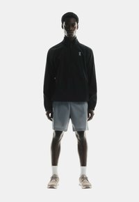 Black zip-up jacket with high collar, light gray shorts, and beige running shoes. Features logo accents and reflective elements.