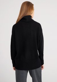 Pull en maille noir avec col roulé haut, poignets et ourlet côtelés. Le tissu semble doux et texturé, porté avec un pantalon gris.