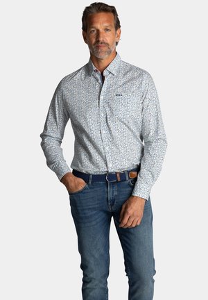 Wit overhemd met blauw en geel patroon; lange mouwen; gecombineerd met blauwe jeans en een marineblauwe riem met bruine accenten.