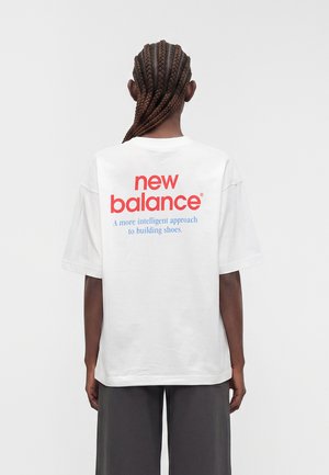 Person med flettet hår iført en hvid New Balance t-shirt med rød og blå logo og tekst på bagsiden, står mod en ensfarvet baggrund.
