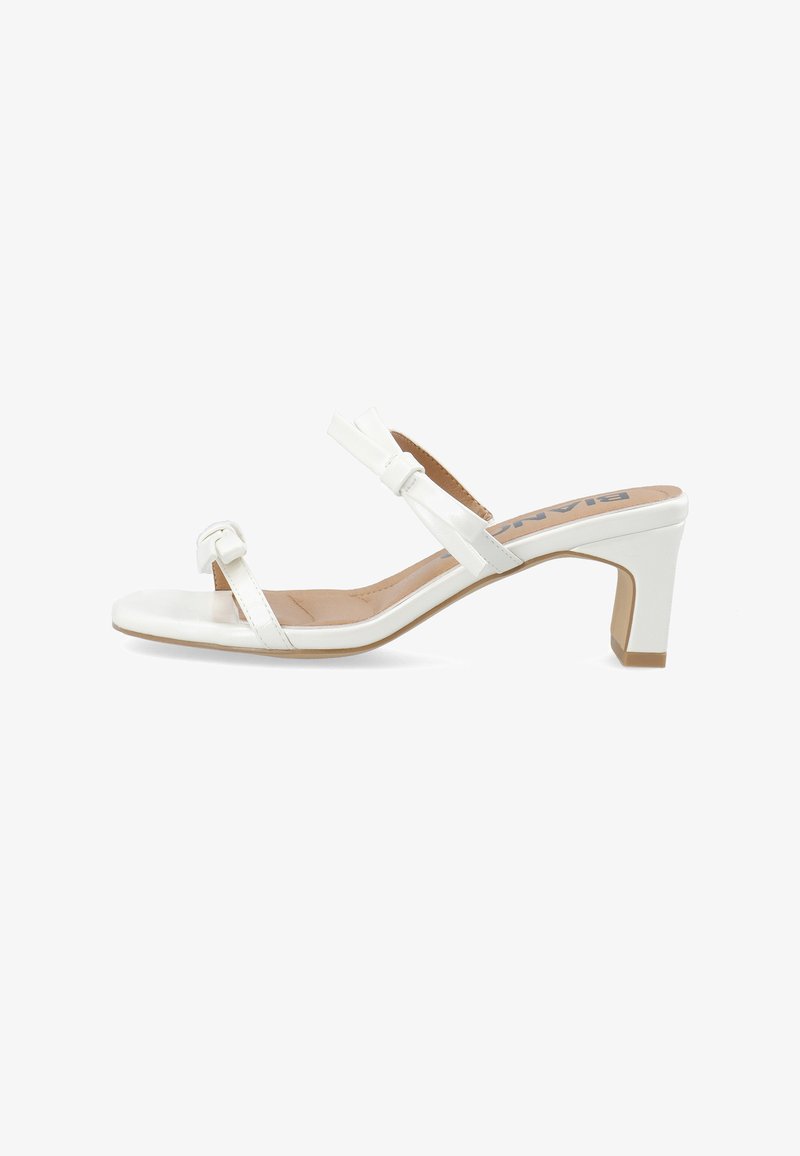 Sandale blanche à talon moyen avec deux brides ornées de petits nœuds, semelle intérieure beige et talon bloc sur un fond blanc uni.