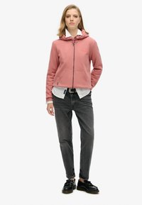 Sudadera rosa con cremallera y capucha, con una textura sólida. Combinada con jeans gris oscuro y zapatos negros, estilizada con una camiseta blanca debajo.