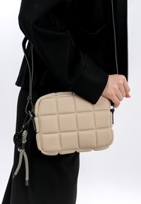 Borsa a tracolla beige trapuntata con forma rettangolare, dotata di una tracolla nera, superficie testurizzata e decorazione con nappina.