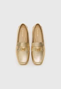 Gold loafers with a textured finish, featuring a decorative band and logo accent. Rounded toe and flat sole design.