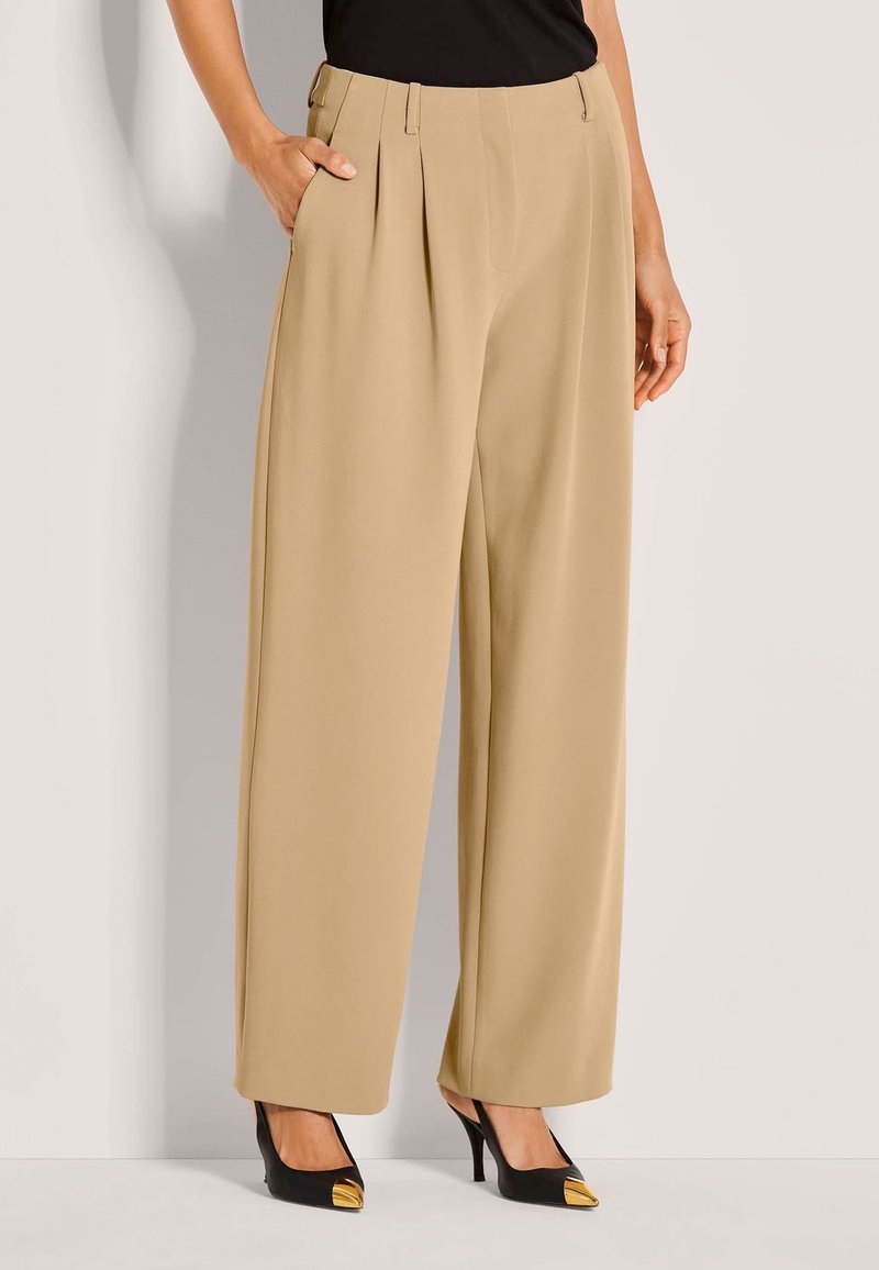 Vrouw in beige high-waisted wijde broek met plooien, zwarte hoge hakken met gouden metallic puntjes, en een zwarte top, staande met hand in de zak.