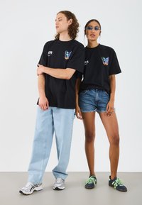 Black graphic t-shirts feature colorful butterfly designs. The male model wears light blue jeans and gray sneakers, while the female wears denim shorts and dark sneakers.