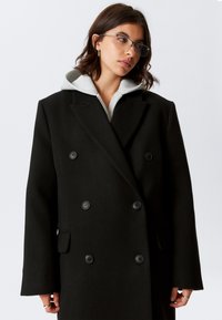 Melnas dubultpogātas virsjaka no teksturētas auduma, ar lielām melnām pogām un atlokāmu apkakli, valkāta virs pelēka kapučveida džempera.