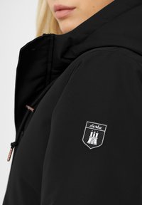 Černá bunda s kapucí, která má výraznou nášivku s designem hradu na levém rukávu a texturovaný povrch látky.