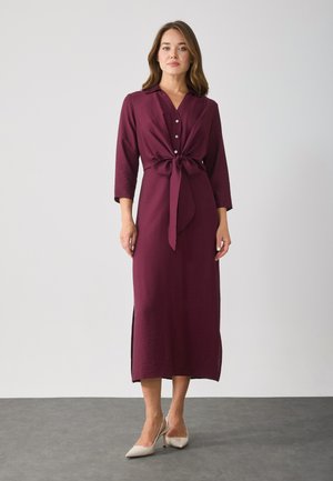 Burgundinvärinen mekko, jossa on vyö, nappilista ja 3/4-hihat. Sisältää sivureikiä ja teksturoidun kankaan. Yhdistetty vaaleisiin korkokenkiin.
