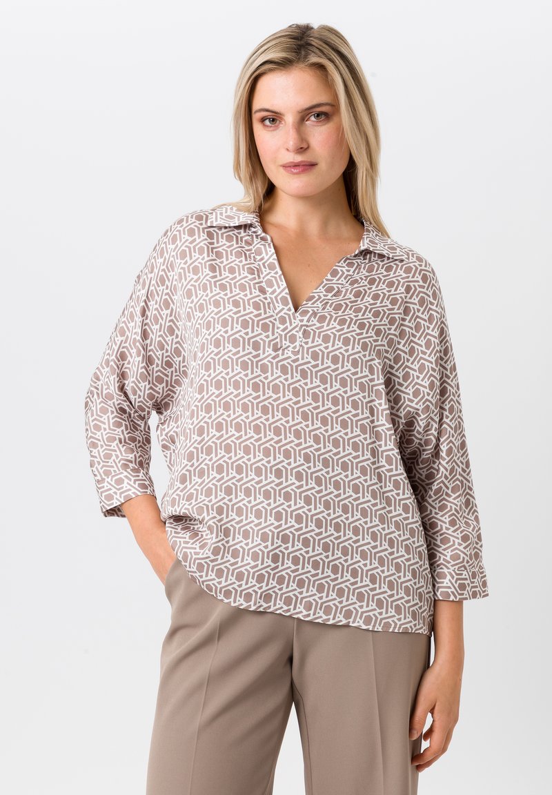 Leichte, beige Bluse mit geometrischem weißen Muster, Kragen und drei Viertelärmeln. Kombiniert mit taillierten beigefarbenen Hosen.