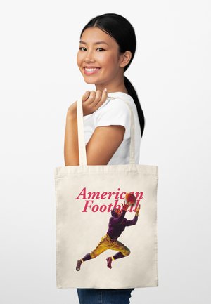 Mladá žena se usmívá a drží béžovou tašku na rameno s ilustrací hráče amerického fotbalu, jak chytá míč.