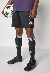 Sort atletiske shorts med hvidt Adidas-logo, sorte sokker med hvide striber og sorte fodboldstøvler, der holder en mønstret fodbold.