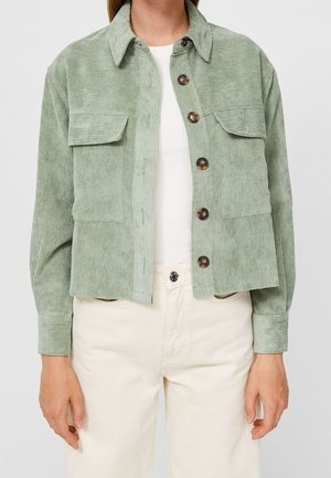 Personne portant une veste boutonnée en velours côtelé vert clair sur un haut blanc et un pantalon crème, montrée du cou aux hanches.