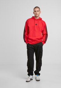 Röd huvtröja med en svart Adidas-logga, med svarta ränder på ärmarna, tillsammans med svarta cargo-byxor och vita sneakers.