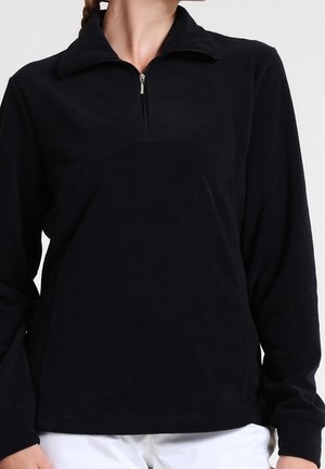 Woman wearing a black long-sleeve fleece pullover with a short zipper at the neck and white pants, against a plain background.