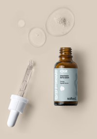 Bottiglia di vetro marrone con un'etichetta grigia che presenta del testo bianco. Include un contagocce con un tappo bianco. Intorno ci sono delle gocce di siero trasparenti su una superficie beige.
