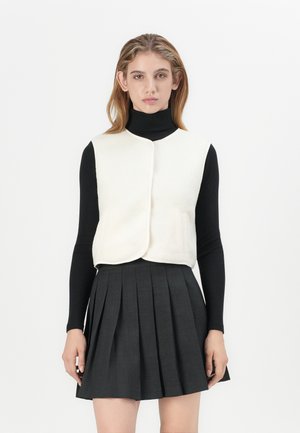 Kremfarget faux-fur vest med rund hals, frontknappeåpning og to sidelommer, båret over en svart turtleneck og en grå plisserte skjørt.