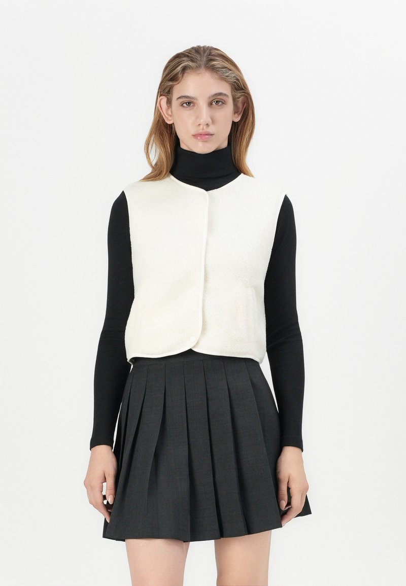 Cremfarbene Kunstfellweste mit rundem Halsausschnitt, knopflosem Verschluss vorne und zwei Seitentaschen, getragen über einem schwarzen Rollkragenpullover und einem grauen Faltenrock.