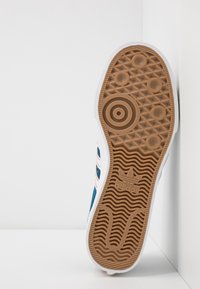 Sole bílé tenisky Adidas s hnědou gumovou podrážkou, která obsahuje kruhové a vlnité vzory, opřená o bílou rohovou zeď.