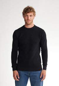 Black, textured crewneck sweater with long sleeves. Ribbed cuffs and hem. Worn with blue denim jeans. No visible logos or patterns.