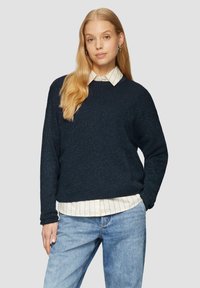 Marineblauwe geribbelde gebreide trui met een ontspannen pasvorm, voorzien van een gestructureerd oppervlak. Gelaagd over een witte gestreepte overhemd met kraag, gecombineerd met lichtblauwe jeans.