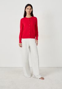 Cardigan rojo con escote redondo y cierre de botones, combinado con pantalones de pierna ancha en gris claro. Textura suave con detalles acanalados en los puños.