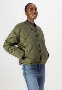 Doudoune vert olive avec un motif matelassé en losanges, fermeture éclair à l'avant et poches latérales. Portée avec un jean bleu, mettant en valeur un look décontracté.
