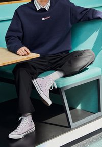 Sneaker alte in tela lavanda chiaro con lacci bianchi, con un'etichetta logo sul lato esterno, indossati con pantaloni neri e una felpa blu navy.