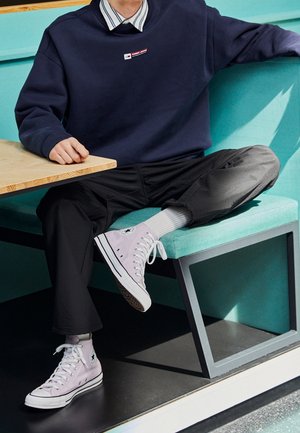 Zapatillas altas de lona color lavanda claro con cordones blancos, con un parche de logo en el lado exterior, combinadas con pantalones negros y un sudadera azul marino.