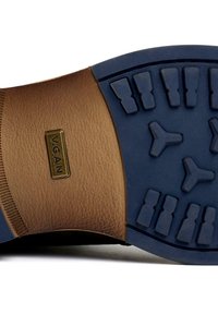Brown vegan leather sole with textured finish, featuring a blue rubber outsole, and embossed branding on a rectangular accent.